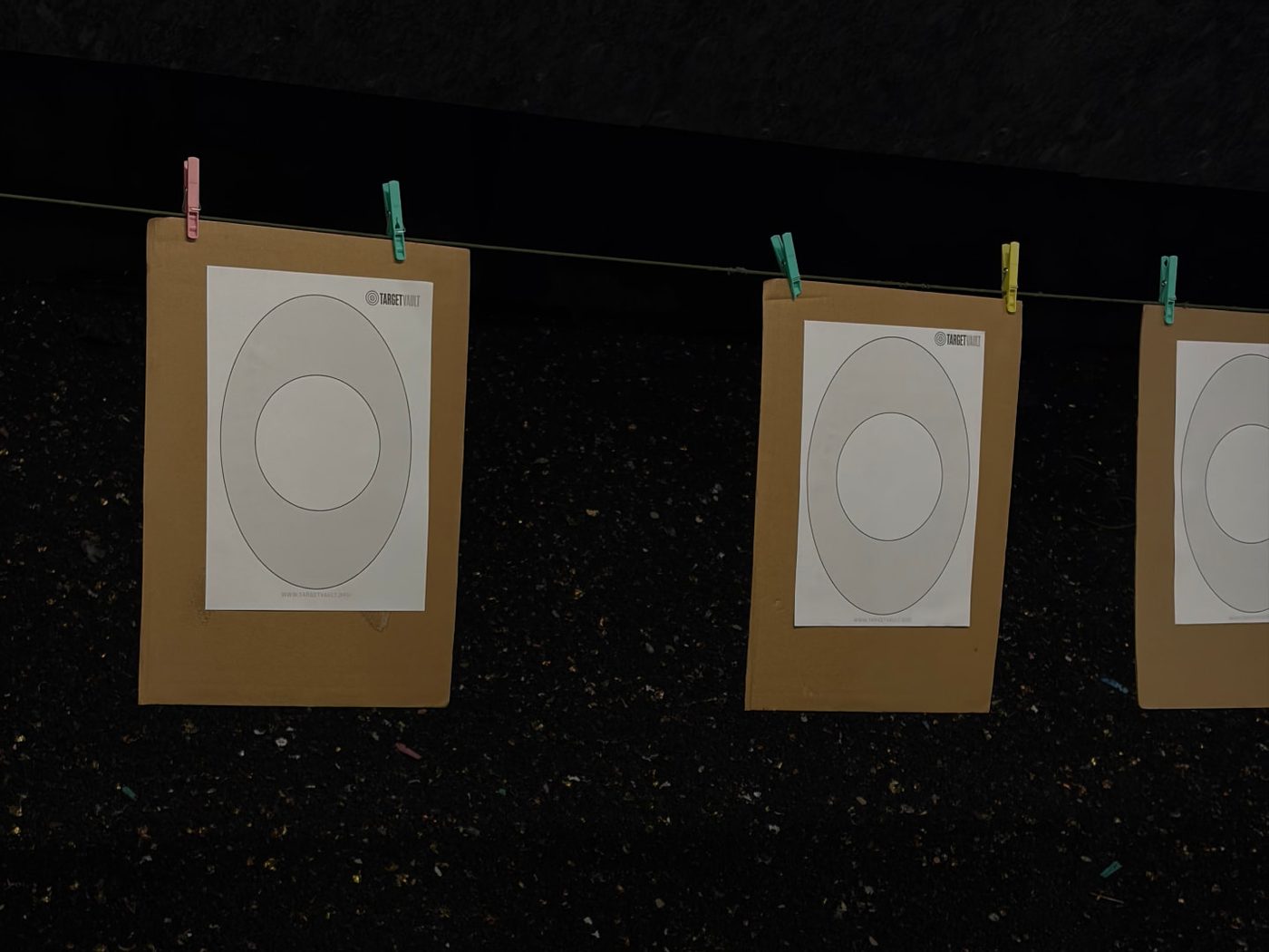 Three printed TargetVault targets clipped to cardboard backers on a line at an indoor range
